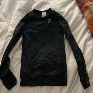 Svart/ mörkgrå långärmad träningströja Gymshark - Svart/ mörkgrå  långärmad träningströja från Gymshark med diskret mönstrad struktur och tumhål vid ärmsluten. Tight passform