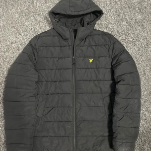 Svart pufferjacka från Lyle & Scott - Svart pufferjacka från Lyle & Scott med huva och dragkedja framtill. Jackan har det klassiska gula fågel-loggan på bröstet och är quiltad med horisontella sömmar. Tillverkad i syntetmaterial och har två sidofickor. Perfekt för kyliga dagar.
