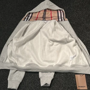 Grå Burberry hoodie med klassiskt foder - Snygg grå hoodie från Burberry i bomull med den ikoniska rutiga insidan av huvan. Tröjan har långa ärmar, ribbade muddar och en stor huva med snörning. Perfekt för dig som vill ha en stilren och lyxig streetwear-look.