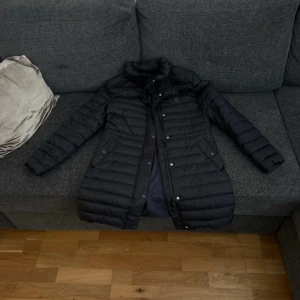 Svart lång dunjacka med dragkedja - Snygg svart lång dunjacka med quiltad design och hög krage. Jackan har både dragkedja och knappar framtill samt två fickor med lock. Perfekt för kyliga dagar och ger en stilren look.