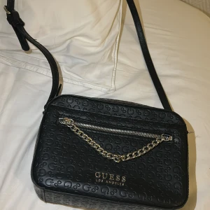 Svart axelväska från Guess med kedja - Snygg svart axelväska från Guess med präglat G-mönster över hela väskan. Väskan har en silverfärgad kedjedetalj framtill, dragkedja upptill och justerbar axelrem. Perfekt storlek för det viktigaste och stilren design med Guess-logga i guld. Jag fick denna i present men den har tyvärr inte kommit till användning.