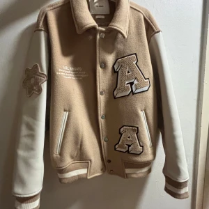 Beige bomberjacka Axel Arigato - Snygg beige bomberjacka från Axel Arigato med vita ärmar i skinn och stora broderade bokstäver på bröstet och ryggen. Jackan har ribbade muddar, knappar framtill och coola detaljer som patch på ärmen.  Köptes för två år sen för 2800 på rea ord. Pris var 3500 säljer för 1000kr -Använd seriöst max 10 gånger, helt i nyskick 