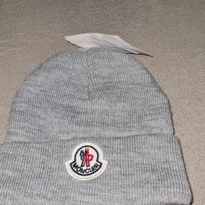 Grå Moncler mössa i stickad ull - Snygg grå mössa från Moncler i ribbstickad ull. Klassisk modell med uppvikt kant och broderad Moncler-logga framtill i vitt, blått och rött. Perfekt accessoar för kyliga dagar och en stilren detalj till din outfit.
