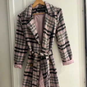 Rutig rosa kappa från Missguided - Säljer en snygg rutig kappa från Missguided i rosa, grått och svart. Jackan har bälte i midjan, stora slag och knappar framtill. Mjuk och lite fluffig yta, helfodrad i ljusrosa tyg. Perfekt för dig som vill sticka ut i höst och vinter!