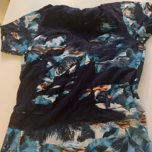 Blå mönstrad t-shirt från Kenzo - Snygg t-shirt från Kenzo i storlek L med coolt blått och svart mönster som påminner om tropiska blad och hav. T-shirten har korta ärmar och är gjord i mjuk bomull. Perfekt för dig som vill sticka ut med en unik look.
