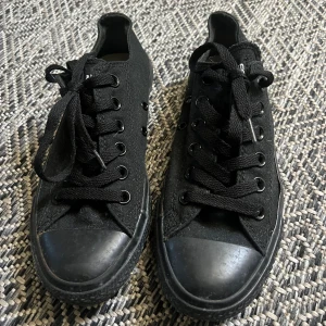 Svarta Converse - Svarta låga sneakers från Converse, rund tå och platt sula. Skorna har snörning med svarta skosnören och är tillverkade i textilmaterial. 