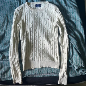 Vit kabelstickad tröja från Gant XS - Vit kabelstickad tröja från Gant i stretchig bomull. Klassisk rund halsringning, långärmad modell och broderad Gant-logga på bröstet. Snygg ribbad kant vid ärmslut och nederkant. Perfekt för en clean och tidlös look.