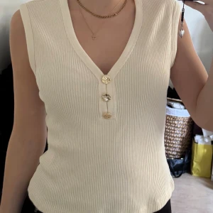 Ribbad vit linnetopp med knappar - Snygg ribbad linnetopp från H&M i vit färg med v-ringning och tre dekorativa guldfärgade knappar framtill. Toppen är ärmlös och har en slim passform som sitter snyggt på kroppen. Perfekt för att skapa en clean och fräsch look.