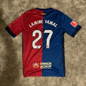 Lamine Yamal #27 debut tröja Nike S - Säljer en FC Barcelona matchtröja med Lamine Yamal och nummer 27 på ryggen. Tröjan är från Nike med Dri-FIT material, har korta ärmar och är delad i röd och blå färg med gula detaljer. Spotify-logga på framsidan och UNHCR ACNUR på baksidan.