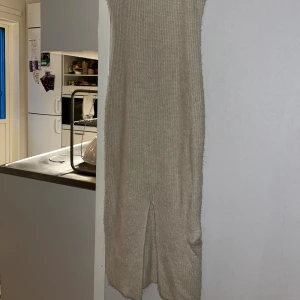 Stickad beige långklänning med slits - Beige stickad långklänning med smala axelband och v-ringning. Klänningen har en tight passform och en snygg slits framtill. Perfekt för dig som gillar en enkel men trendig look med stickad känsla.