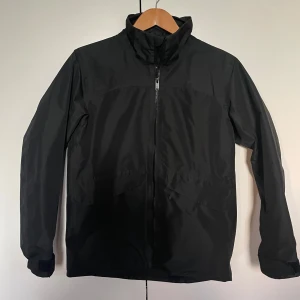 Svart vindjacka från Everest - Svart vindjacka från Everest med hög krage och hel dragkedja framtill. Jackan har långa ärmar, justerbara muddar och en diskret reflexdetalj baktill. Tillverkad i lätt och smidig polyester, perfekt för blåsiga dagar. Storlek 158/164