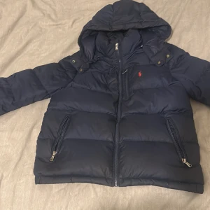 Mörkblå dunjacka från Ralph Lauren - Snygg mörkblå dunjacka från Ralph Lauren med huva och dragkedjor på fickorna. Jackan har det klassiska röda Polo-loggan broderad på bröstet och är vadderad för extra värme. Perfekt för kalla dagar och har en stilren look. Ny pris 3200 mitt pris 1900kr säljer jag pågrund av att jag har väckt ur den jag köpte den förra vintern.