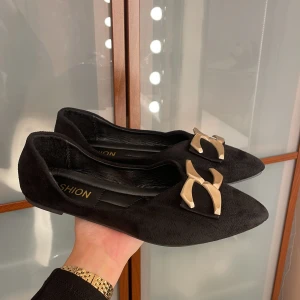 Svarta ballerinaskor med gulddetalj - Snygga svarta ballerinaskor med spetsig tå och mjukt mockaliknande material. På ovansidan sitter en stor guldfärgad metallrosett som ger skorna en cool och trendig look. Perfekt för dig som gillar stilrena detaljer och vill sticka ut lite extra.