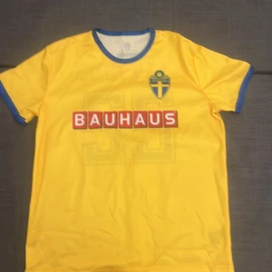 Svenska landslagets gul fotbollströja - Gul fotbollströja från svenska landslaget med blå detaljer och Bauhaus-logga på både fram- och baksida. Nummer 22 i blått på ryggen och landslagsemblem på bröstet. Tillverkad i lätt och ventilerande polyester, perfekt för match eller träning.