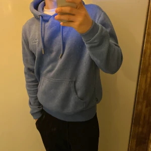 Blå hoodie från Superdry med huva - Säljer en blå hoodie från Superdry i storlek M. Tröjan har en klassisk huva med snörning, magficka och ribbade muddar vid ärmslut och nederkant. Mjuk insida i fleece och diskret Superdry-logga på bröstet. Perfekt för chill dagar.