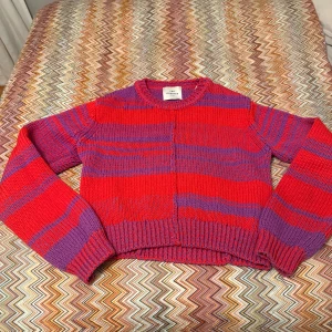 Randig stickad tröja med öppen rygg - Säljer en färgstark stickad tröja från Les Coyotes i rött och lila med breda ränder. Tröjan har lång ärm, ribbade muddar och en unik öppen ryggdetalj som gör den extra cool. Perfekt för dig som vill sticka ut med din stil!