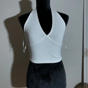 Vit ribbad halterneck topp - Säljer då den inte passar längre. Toppen är insydd i nacken. Längd ca 40cm. Längd runt halsen ca 50cm. Pris: 10kr 