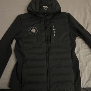 Svart dunjacka Sail Racing XS - Svart dunjacka från Sail Racing i storlek XS med huva och snygga logodetaljer på bröstet och ärmen. Den är dock sönder vid handleden tyvärr, men den kan enkelt bli sydd.