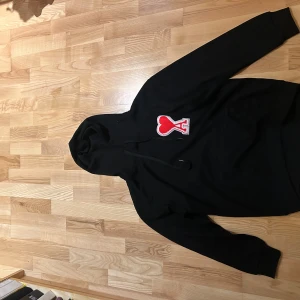 Svart hoodie från Ami med rött hjärta - Svart hoodie från Ami Alexandre Mattiussi med stor röd hjärtpatch och bokstaven A på bröstet. Klassisk huva med snörning och känguruficka framtill. Tillverkad i mjuk bomull, perfekt för en chill och stilren look.