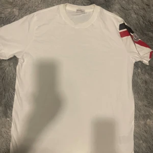 Vit Moncler t-shirt med logga - Snygg vit t-shirt från Moncler med klassisk rund hals och korta ärmar. Ena ärmen har coola ränder i svart och rött samt Moncler-logga som detalj. Tillverkad i mjuk bomull som känns skön mot huden. Perfekt för dig som gillar stilrena plagg med en twist.