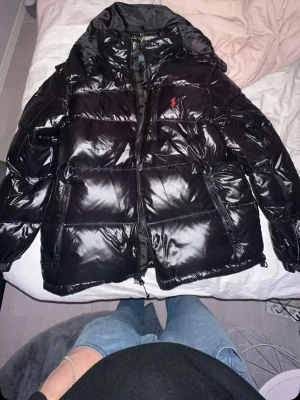 Ralph Lauren jacka  - Ej hugget i sten Säljer en svart, glansig pufferjacka från Polo Ralph Lauren med röd logga på bröstet. Jackan har huva, dragkedja framtill och två sidofickor med dragkedja. Perfekt för kalla dagar och riktigt snygg stil.