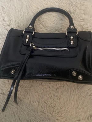 Svart handväska med dragkedja - Snygg svart handväska i skinn med silverfärgade nitar och detaljer. Väskan har ett kort handtag och en dragkedja framtill med dekorativa remmar. Perfekt storlek för det viktigaste och har en cool, stilren look.