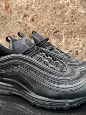 Nike Air Max 97 Triple Black sneakers 40,5 - Mycket fint skick, anvönda några få gånger Nike Air Max 97 Triple Black sneakers med vågiga paneler och synlig Air-sula. Hela skon är svart med mesh och syntetmaterial, samt diskreta Nike-loggor på sidan och hälen. Klassisk snörning och rund tå. Perfekt för dig som gillar streetwear och sportig stil.