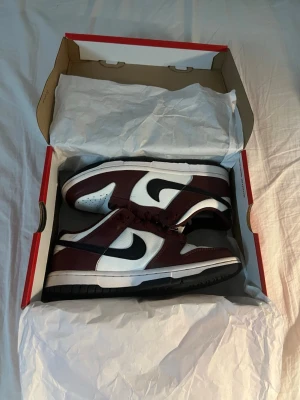 Nike Dunk Low vit och vinröd - Nike Dunk Low sneakers i vit och vinröd färg med svarta detaljer och klassisk svart swoosh. Skorna har snörning, perforerad tå och platt sula. Ovandelen är i skinn och de har en låg siluett som ger en clean och stilren vibe.