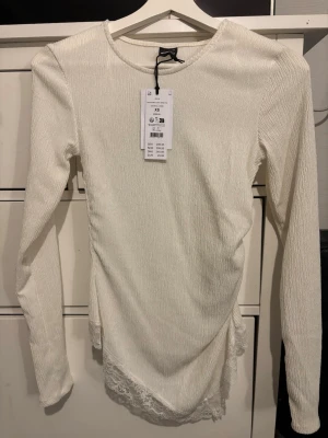 Vit asymmetrisk blus med spets från Gina Tricot - Säljer en vit långärmad blus från Gina Tricot i storlek XS. Toppen har asymmetrisk nederkant med spetsdetaljer och är tillverkad i ett strukturerat syntetmaterial. Rund halsringning och figurnära passform. Perfekt för dig som gillar unika detaljer.