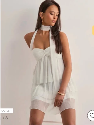 Vit volangklänning från Nelly - Supersöt vit kort klänning från Nelly med flera lager volanger och knytband i halsen. Klänningen är ärmlös och har en romantisk känsla med sitt tunna, luftiga tyg. Perfekt för sommarens alla tillfällen och ger en riktigt fräsch look.