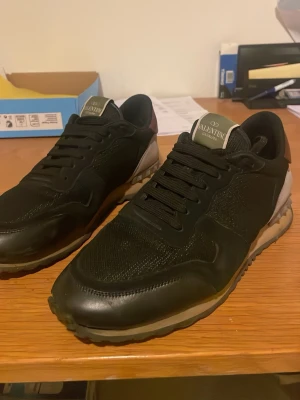 Valentino Garavani Rockrunner sneakers - Säljer ett par svarta Valentino Garavani Rockrunner sneakers med coola detaljer i mocka, skinn och mesh. Skorna har svarta snören, beige och grå inslag på hälen samt den klassiska nitar-detaljen bak. Snygg och sportig siluett som passar till många outfits. Kan gå ner i pris vid snabb affär!