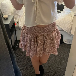 Blommig kortkjol med volang - Kjol i rosa toner med volangdetaljer. Kjolen har en elastisk midja. Lite liten i storleken. Köpt på plick men aldrig använt själv💕