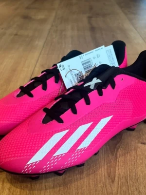 Adidas rosa fotbollsskor  - Säljer ett par grymt snygga fotbollsskor från Adidas i stark rosa med svarta och vita detaljer. Skorna har snörning, rund tå och en platt sula med svarta dobbar för extra grepp på planen. Ovandelen är tillverkad i syntetmaterial med mönstrad struktur. Helt nya