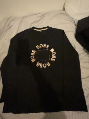 Svart långärmad tröja från Hugo Boss - Svart långärmad tröja från Hugo Boss med coolt tryck i guld på bröstet där det står 'BOSS' i en cirkel. Tröjan har rund hals och är gjord i mjuk bomull, perfekt för dig som gillar stilrena och trendiga plagg med lite extra attityd.