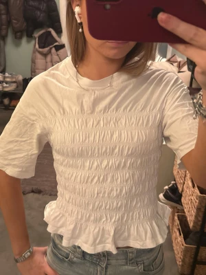 Vit smockad blus från Gina Tricot - Supersöt vit blus från Gina Tricot i storlek S. Toppen har smockad midja, volang nertill och korta ärmar. Perfekt till jeans eller kjol för en fräsch look. Materialet känns mjukt och lätt, och den har en klassisk rund halsringning.