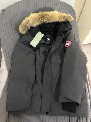 Mörkgrå dunjacka från Canada Goose - Säljer en mörkgrå dunjacka från Canada Goose med fluffig päls på huvan och klassisk logga på ärmen. Jackan är en av den långa moderllerna och det står XL men det passar L 