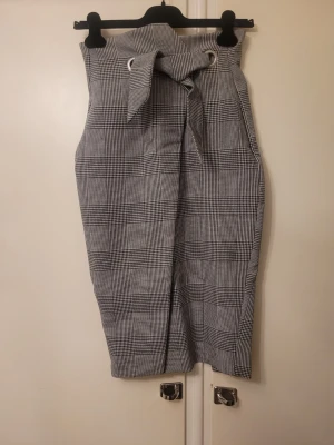 Rutig långkjol med knyt från H&M - Säljer en snygg rutig långkjol från H&M i svart och vitt. Kjolen har ett högt midjeband med knyt och metallöljetter framtill samt en slits bak för extra rörelsefrihet. Perfekt för dig som gillar klassisk stil med en modern twist.