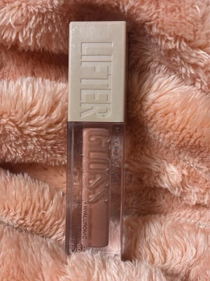 Maybelline Lifter Gloss nude - Maybelline Lifter Gloss i en nude/ljusbrun nyans med hyaluronsyra. Kommer i en genomskinlig fyrkantig plastförpackning med beige lock och stor applikator. Ger glansiga och återfuktade läppar med en mjuk finish. Använd bara 2 gånger