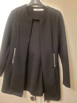 Svart kavajjacka från Vero Moda - Stilren svart kavajjacka från Vero Moda med snygga silverfärgade dragkedjor vid fickorna. Jackan har en rak passform, rund halsringning och är långärmad. Materialet är strukturerat och ger en modern vibe.