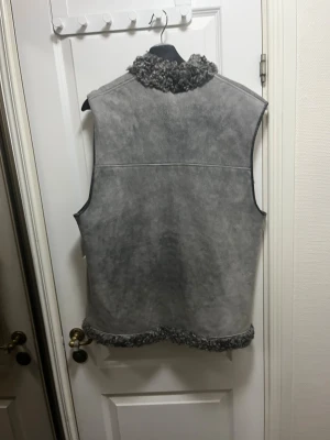 Grå vändbar pälsväst i äkta fårskinn och äkta får päls  - Säljer en vändbar grå pälsväst i fårskinn med Äkta mjuk lockig päls på ena sidan och mockaliknande yta på andra. Västen har två fickor och hög krage, samt pälsdetaljer längs kanterna. Perfekt för dig som vill ha en unik och varm look.