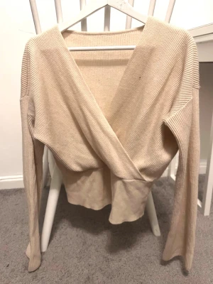 Beige omlott kofta med ribbad struktur - Säljer en beige omlott kofta med ribbad struktur och långärmad design. Koftan har en djup v-ringning och är tillverkad i ett mjukt stickat material som ger en skön och avslappnad känsla. Perfekt för lager-på-lager och passar till många olika stilar.