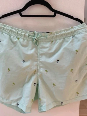 Nikben badshorts med palmmönster - Säljer ett par badshorts med små broderade palmer och parasoller. De har elastisk midja med snörning och en bakficka med lock. Perfekta för sommardagar och strandhäng. Materialet känns som bomull och de har en loose passform.