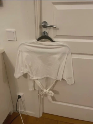 Vit croppad topp med knyt - Säljer en vit croppad topp med knyt i midjan. Toppen har rund hals och korta ärmar, tillverkad i mjukt bomullsmaterial. Perfekt för dig som gillar enkel och stilren design med en twist. Passar till jeans, kjol eller shorts.