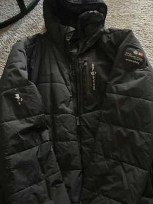 Svart pufferjacka från Sail Racing - Svart pufferjacka från Sail Racing med huva och coola logodetaljer på bröstet och ärmen. Jackan har dragkedja framtill och flera praktiska fickor. Perfekt för kalla dagar när du vill hålla stilen och värmen. Men de har lite färg klumpar på ena ärmen 
