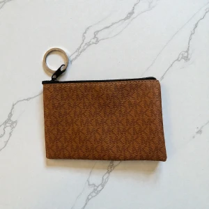 Brun plånbok med dragkedja från Michael Kors - Snygg brun plånbok i skinnimitation med diskret mönster och svart dragkedja. Praktisk ring för nycklar eller att fästa i väskan. Perfekt storlek för kort och småsaker.