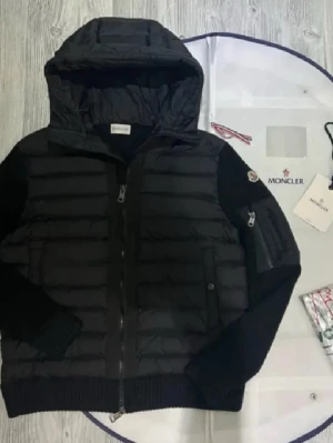Svart pufferjacka från Moncler - Svart pufferjacka från Moncler med huva och ribbade muddar. Jackan har quiltad front, stickade ärmar och dragkedja framtill. Moncler-logga på ärmen och praktisk ficka med dragkedja. Perfekt för kalla dagar och snygg streetstil.