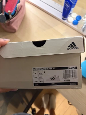 Adidas Grand Court Base 2.0 sneakers - Säljer ett par Adidas Grand Court Base 2.0 sneakers i vitt med klassiska svarta ränder på sidan. Skorna har en stilren och tidlös design med låg profil och platt sula. Perfekta för dig som gillar enkel och sportig stil. Materialet är syntet och textil. Storlek 40.