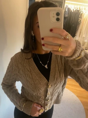 Beige stickad kofta med guldknappar - Säljer en beige stickad kofta med snyggt flätmönster och guldiga knappar framtill. Koftan har v-ringning, ribbade muddar och är croppad i modellen. Perfekt att slänga över en topp för en chill men ändå uppklädd look.