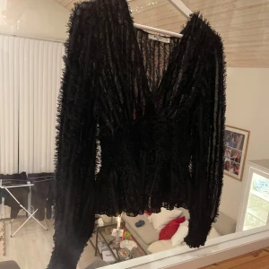 Svart transparent blus från NA-KD - Säljer en svart blus från NA-KD i storlek 38. Blusen är transparent med fluffiga, texturerade ränder och har en djup v-ringning. Långa ärmar och lätt croppad passform ger en cool och trendig look. Perfekt att styla med höga jeans eller kjol.