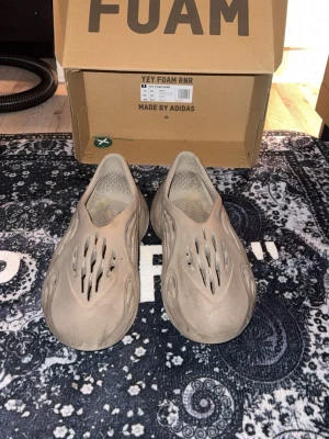 Yeezy Foam Runner - Säljer ett par Adidas Yeezy Foam Runner i beige färg. Skorna har en futuristisk design med många öppna hål och rundad form. Tillverkade i syntetmaterial som är lätt och bekvämt. Perfekta för chill dagar och har en platt sula. Äkta följer med tag som intygar äktheten och självklart boxen och tofflorna 